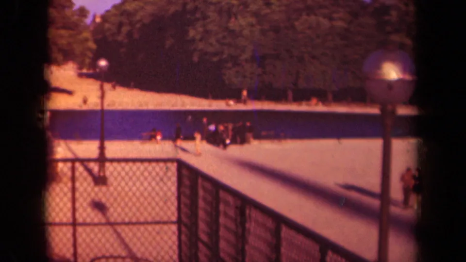 A distant group in a 1939 Paris park gathers beside a blue-tinted water feature, seen past a chain-link fence and lampposts in soft 8mm color.