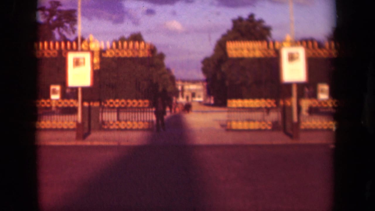 1939 Paris home-movie frame showing an ornate gate, white signboards, trees, and a walkway leading toward a distant building.