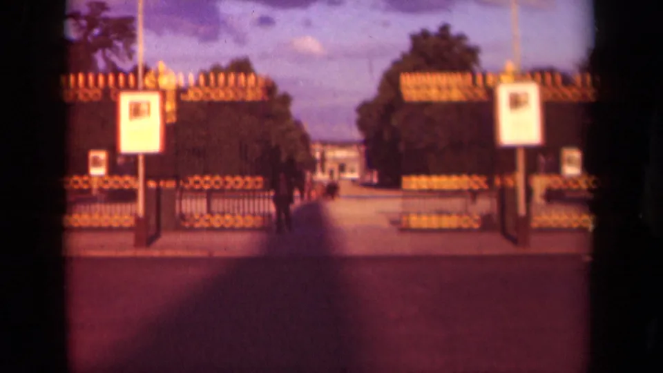 1939 Paris home-movie frame showing an ornate gate, white signboards, trees, and a walkway leading toward a distant building.