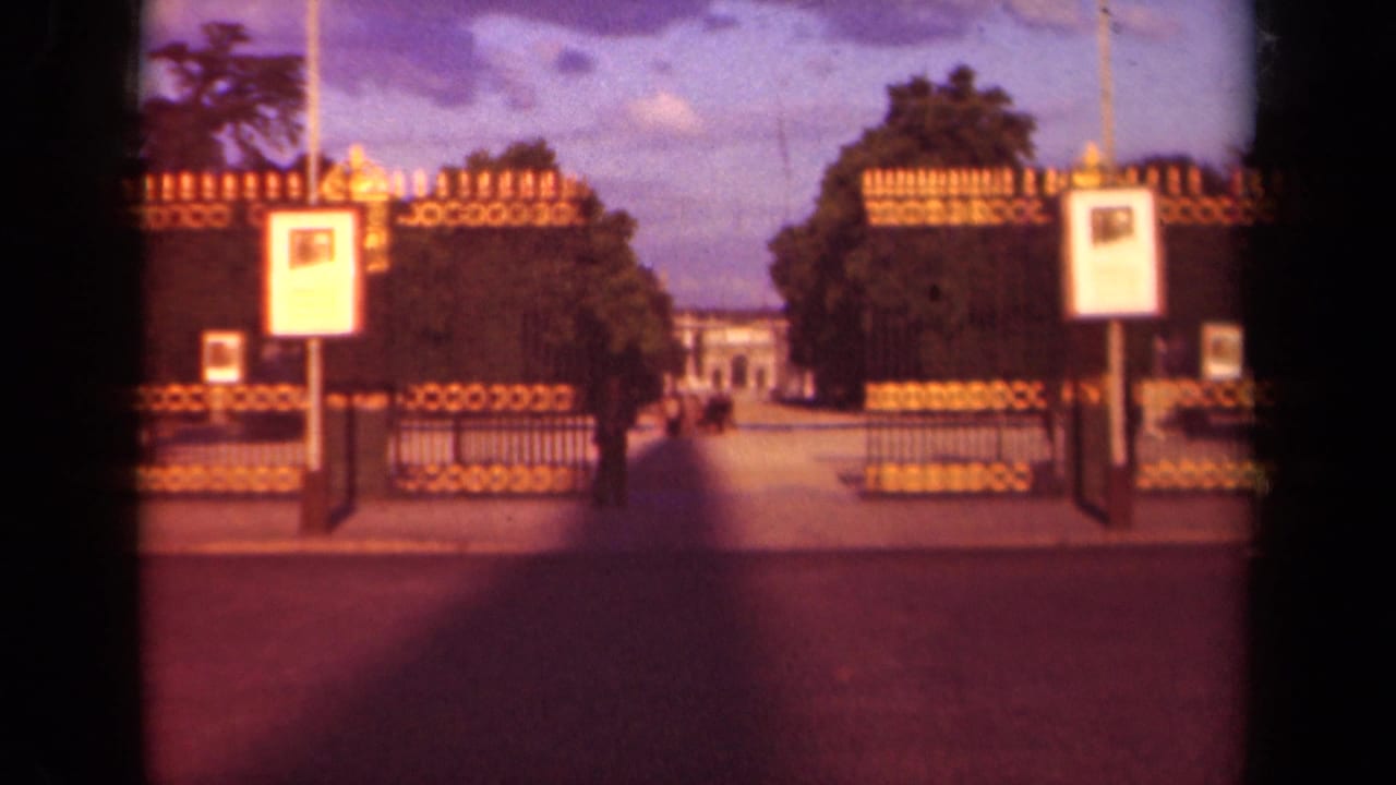 1939 Paris home-movie frame holding on the ornate entrance, with signs in front, trees on both sides, and a distant building centered beyond the gate.