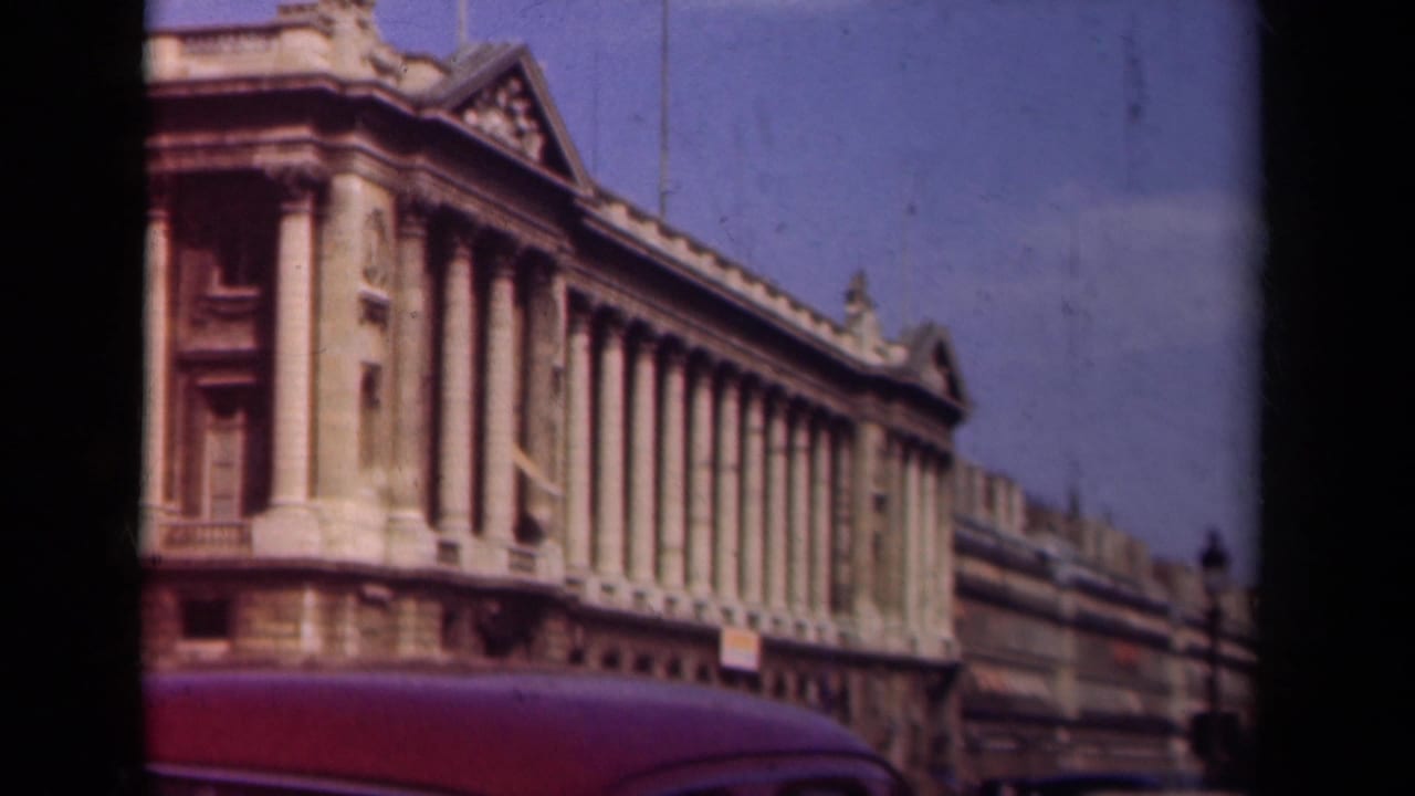 A late-1930s Paris street angle frames a grand columned facade, blue sky, and the top of a red automobile.