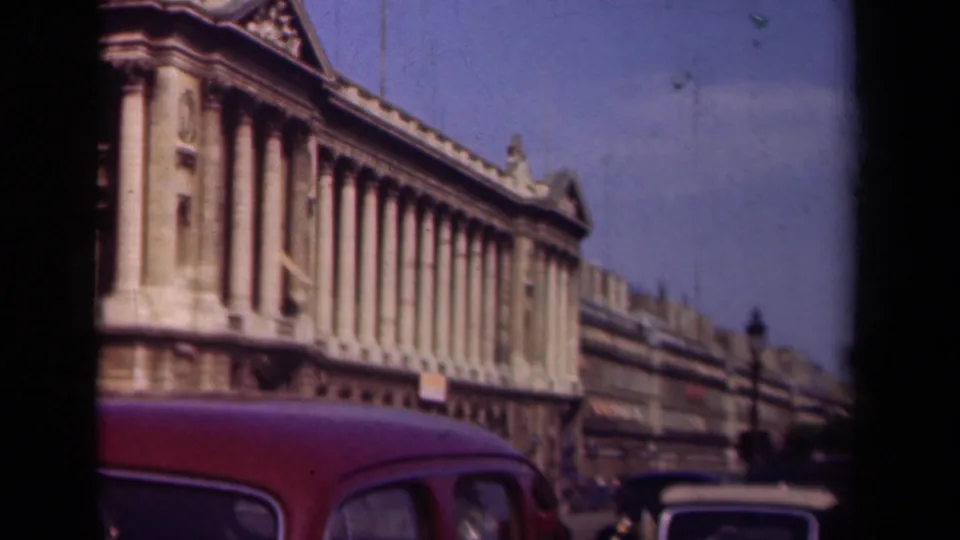 1939 Paris home-movie frame with a grand facade, red foreground car, and street traffic beginning to appear at right.