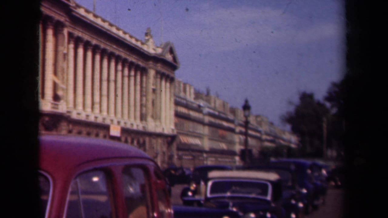 1939 Paris street traffic with a red car at left, dark sedans, lampposts, and a large columned facade in soft 8mm color.