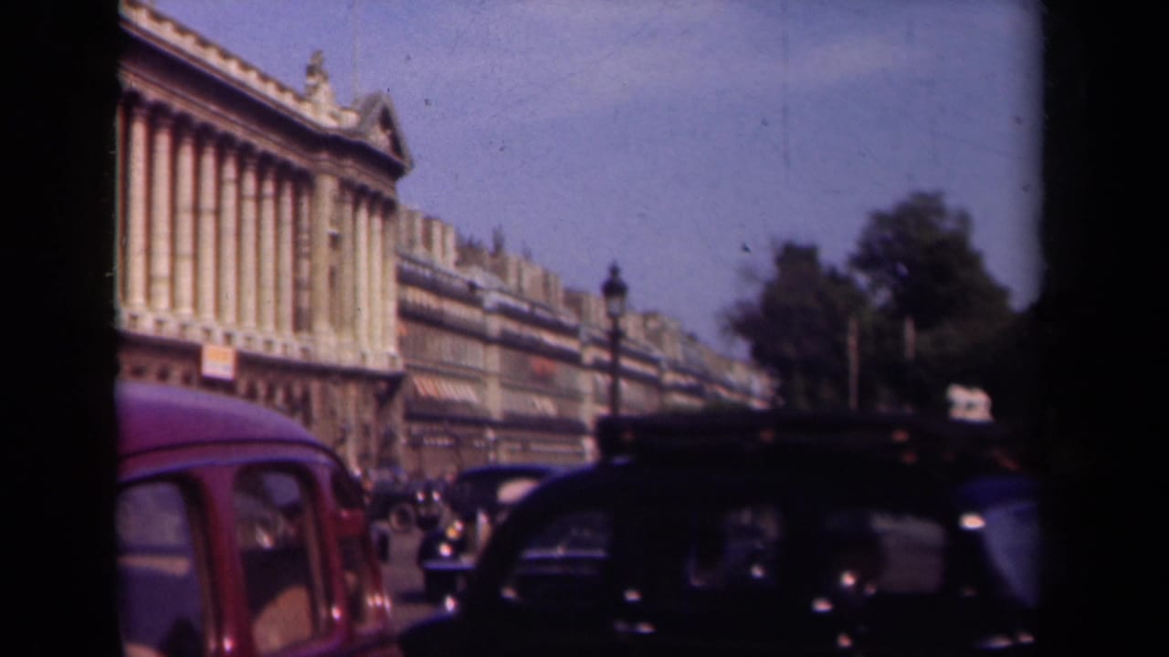1939 Paris traffic scene with dark cars, a red automobile, street lamps, trees, and a long classical facade seen through 8mm frame edges.