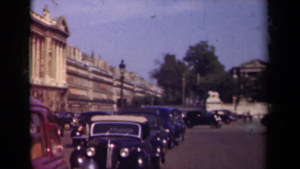 1939 Paris boulevard view with an approaching dark sedan, parked or moving cars, a red car edge, trees, and classical architecture in faded color.