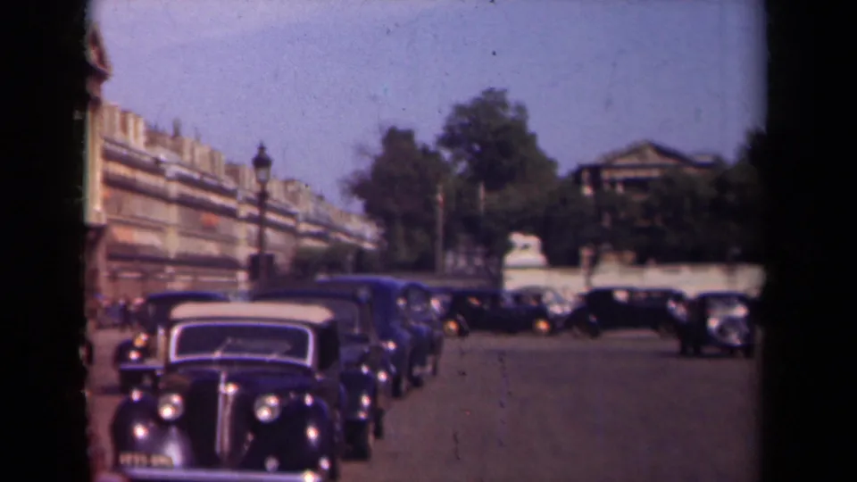1939 Paris street traffic with several dark automobiles, a broad roadway, lampposts, trees, and formal building facades in soft 8mm footage.