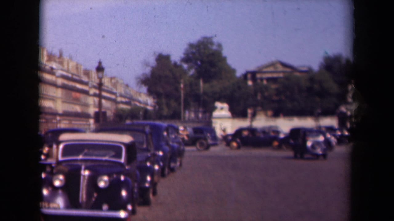 1939 Paris home-movie frame showing rows of dark automobiles along a broad street with trees, apartment facades, and stone gate features beyond.