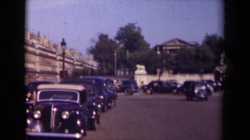 1939 Paris home-movie frame showing rows of dark automobiles along a broad street with trees, apartment facades, and stone gate features beyond.