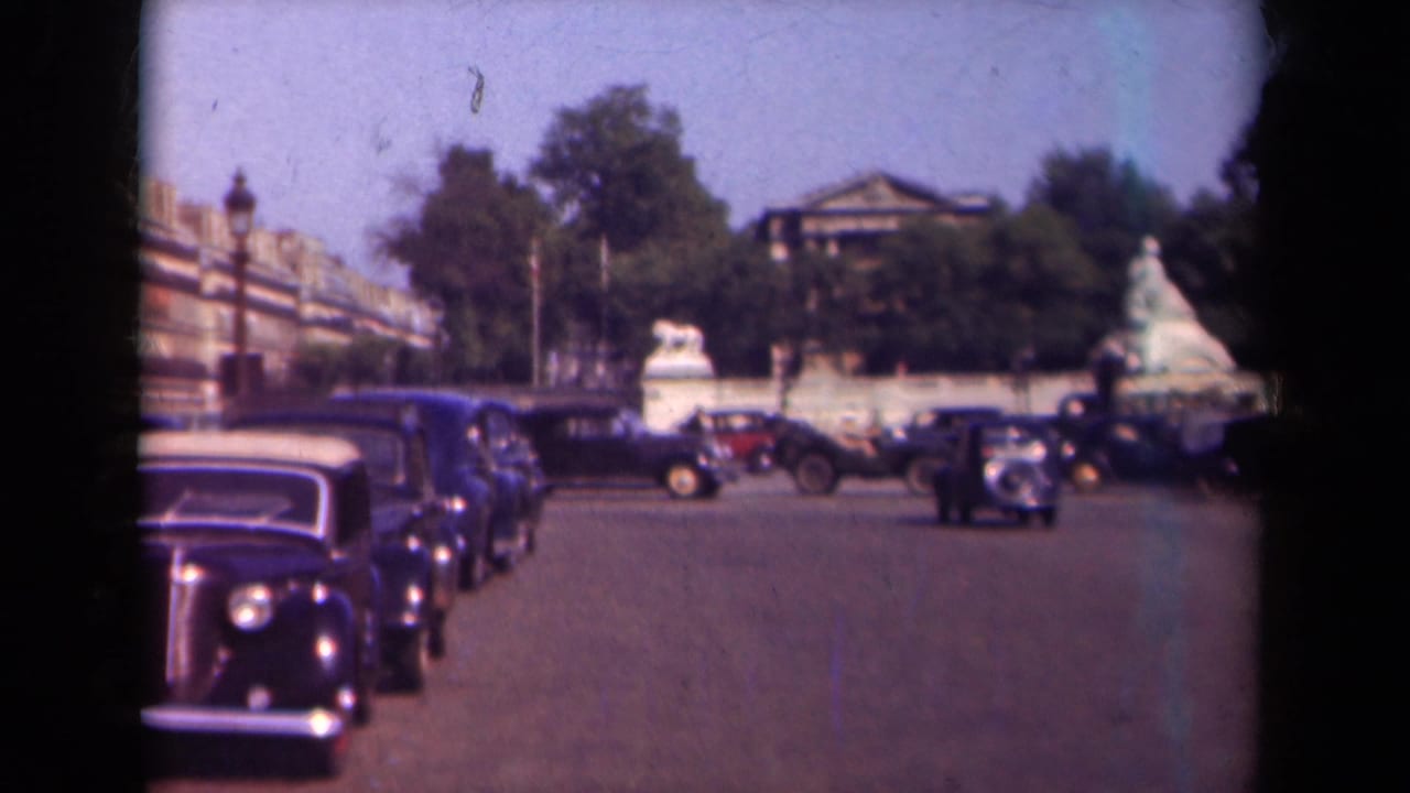 1939 Paris street view with classic automobiles moving through the frame while parked sedans line the road near trees and stone boundary walls.