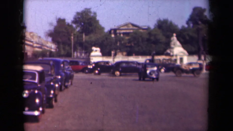1939 Paris 8mm frame showing a line of dark automobiles along a wide street, with leafy trees, stone wall details, and distant buildings behind.