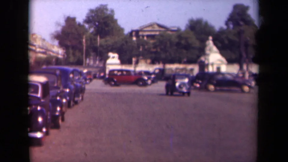 1939 Paris home-movie frame with parked and moving automobiles, including a red vehicle, set against trees, stone walls, and distant urban facades.