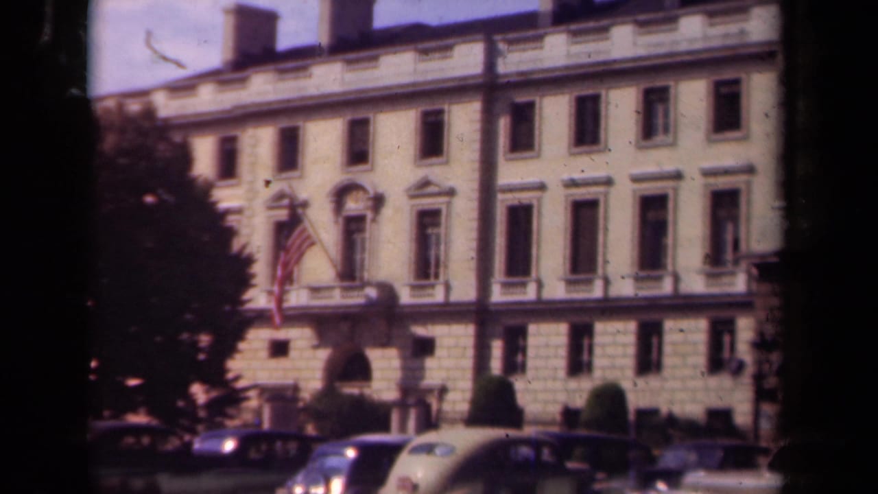 Paris 1939 frame of a classical public building facade, American flag, trimmed greenery, and 1930s automobiles at curbside.