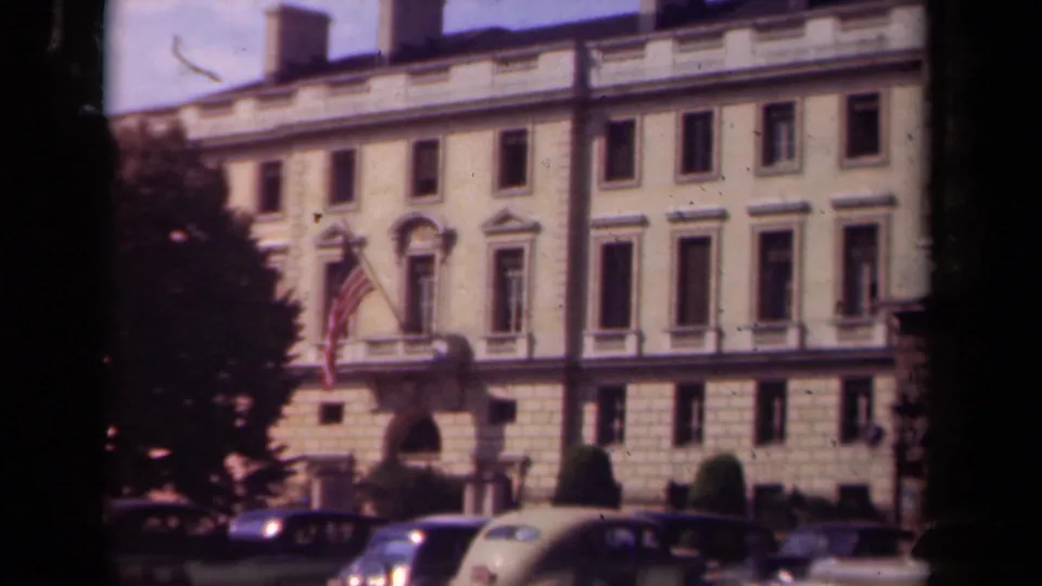 Paris 1939 frame of a classical public building facade, American flag, trimmed greenery, and 1930s automobiles at curbside.