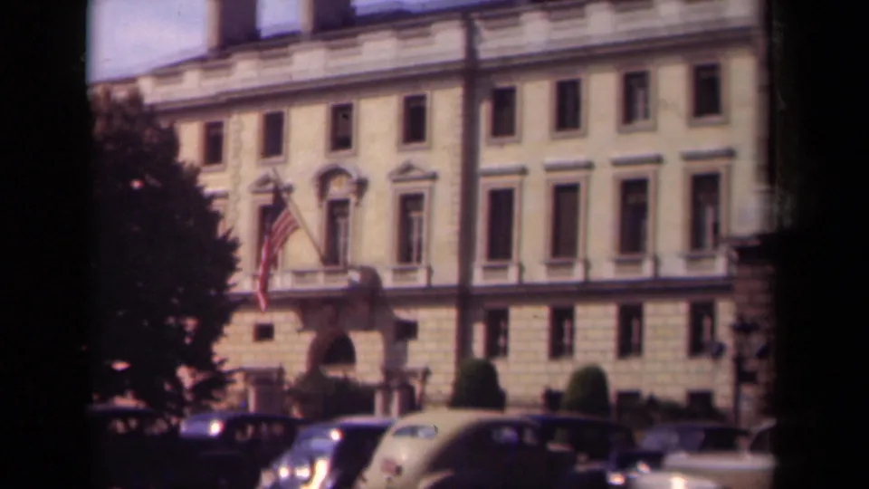 Soft 8mm frame from Paris in 1939 showing the upper facade, American flag, arched doorway, trees, and parked cars below.