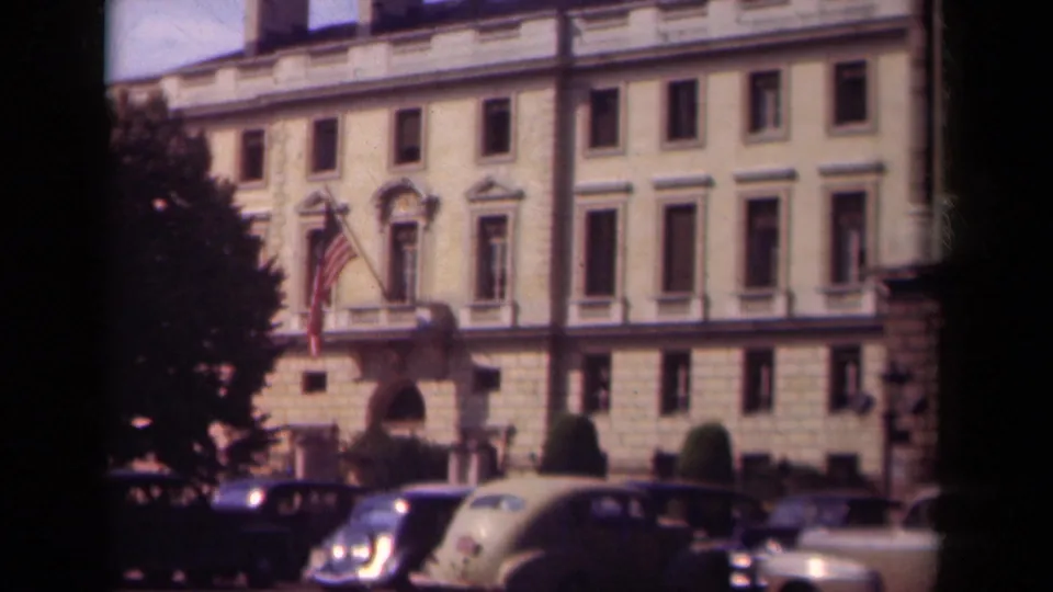 Paris 1939 home-movie frame with a classical stone exterior, American flag near the entrance, parked cars, trees, and visible 8mm frame borders.