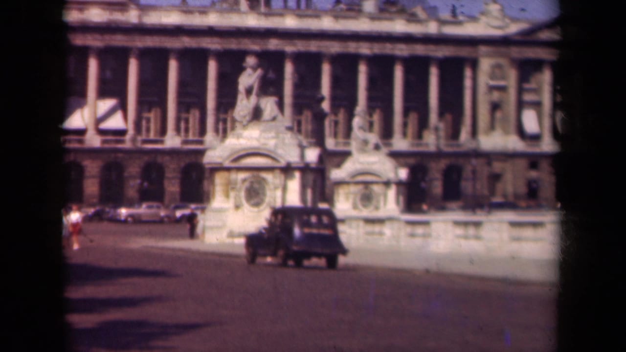 1939 Paris home-movie frame showing a dark car passing before a large neoclassical building with sculptural stonework.