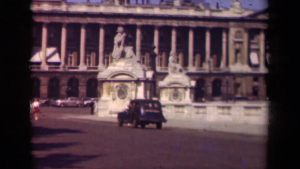 1939 Paris home-movie frame showing a dark car passing before a large neoclassical building with sculptural stonework.