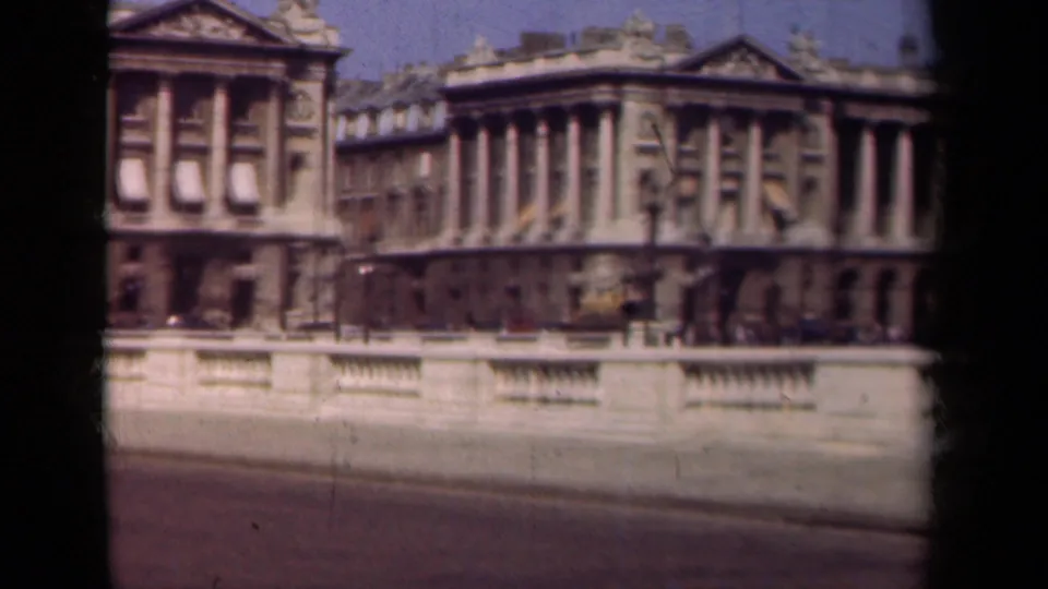 1939 Paris home-movie frame showing a moving view past classical architecture, with columns, roofline sculpture, roadway, and stone railing.