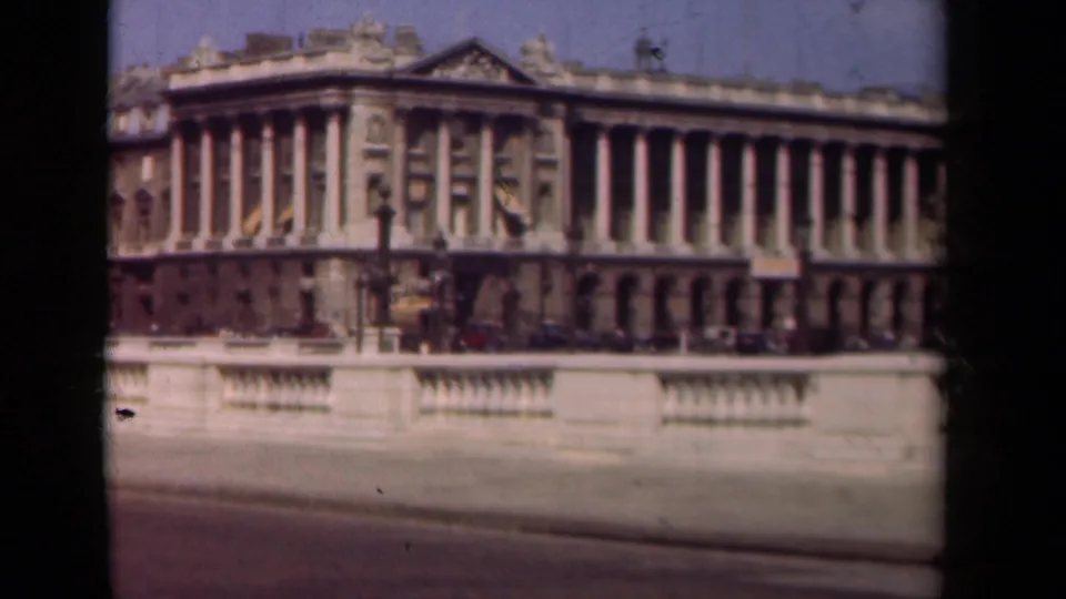 1939 Paris home-movie frame showing a large classical facade with columns, a stone railing, street lamps, and parked cars.