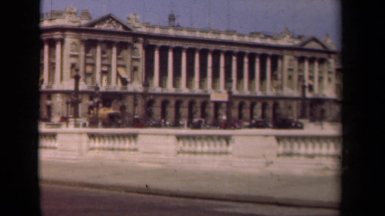 Wide facade with tall columns, ornate roofline, cars, and a stone railing.