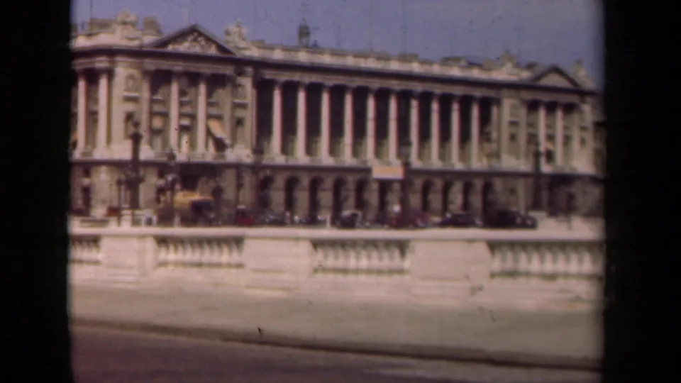1939 Paris frame of a grand colonnaded building with decorative roof details, street lamps, parked cars, and foreground railing.