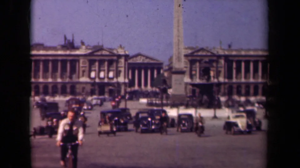 1939 Paris street scene with a cyclist in the foreground, vintage cars in the plaza, and a tall obelisk before classical buildings.