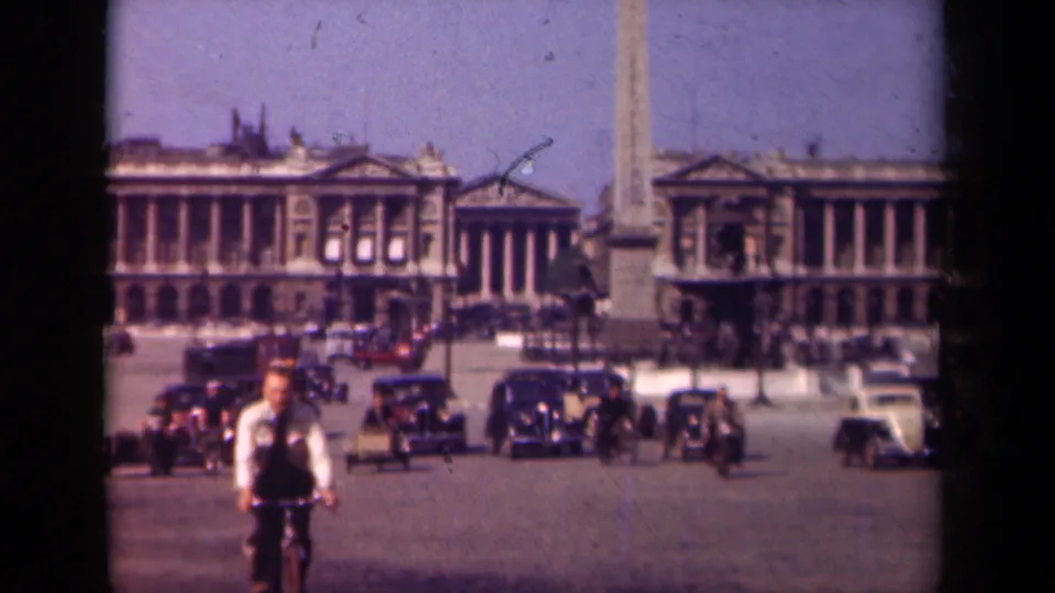 Paris, 1939, showing a cyclist nearing the camera, multiple vintage cars, pedestrians, and an obelisk in a broad urban square.