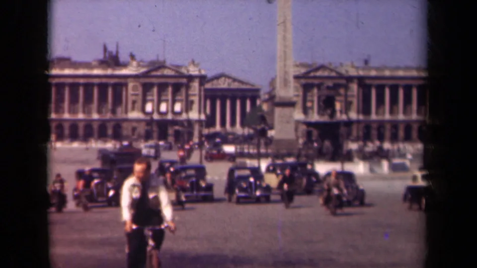 Final 1939 Paris frame with the cyclist close to camera, period automobiles across the plaza, pedestrians, an obelisk, and classical architecture.