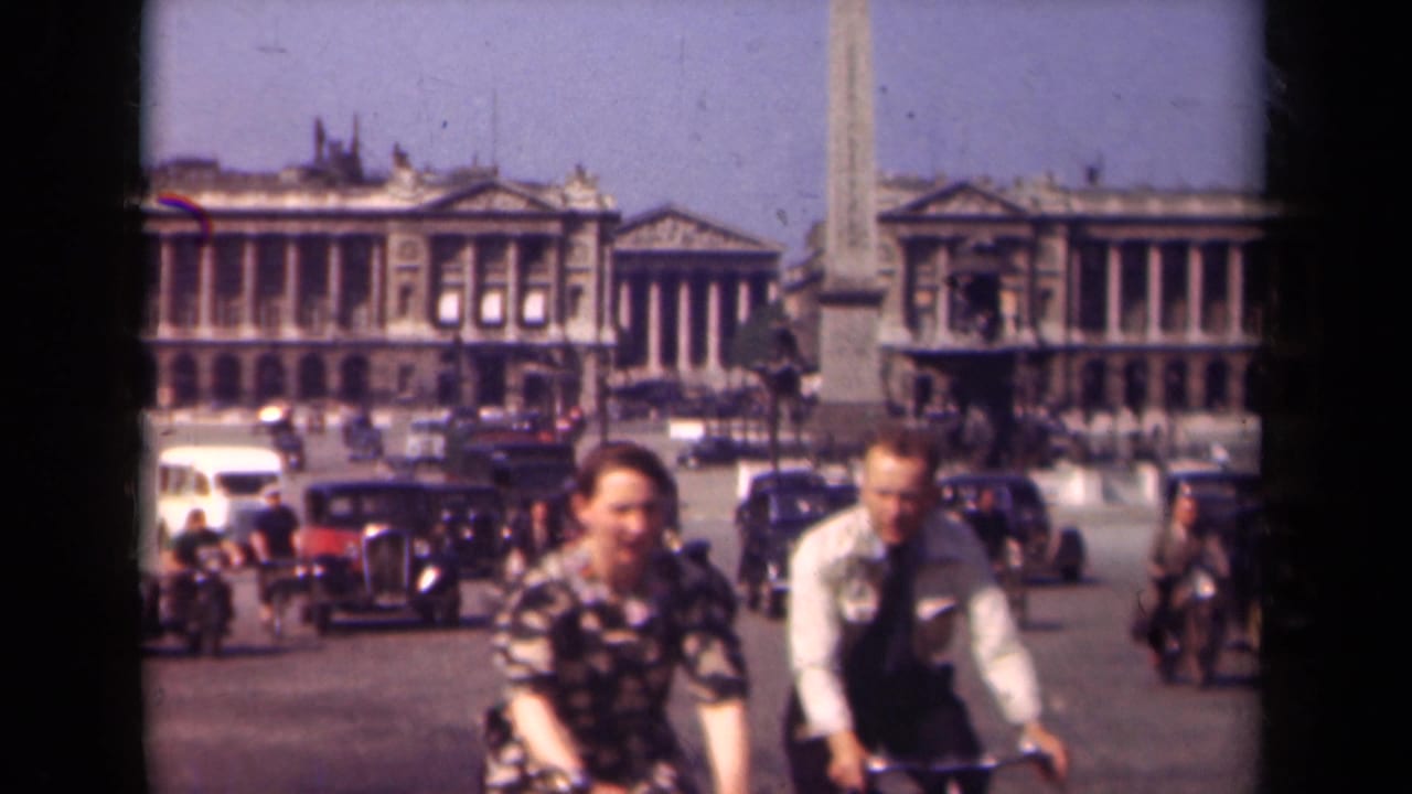 1939 Paris home-movie frame showing cyclists in the foreground, vintage automobiles, pedestrians, an obelisk, and neoclassical buildings across the square.