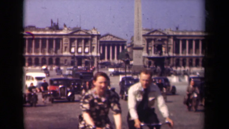 1939 Paris home-movie frame showing cyclists in the foreground, vintage automobiles, pedestrians, an obelisk, and neoclassical buildings across the square.