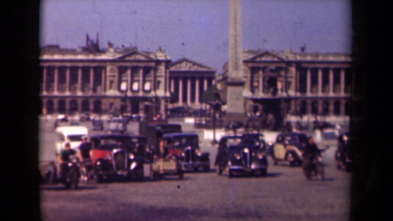 Vintage cars and cyclists move through a broad square with an obelisk and formal buildings in the background.