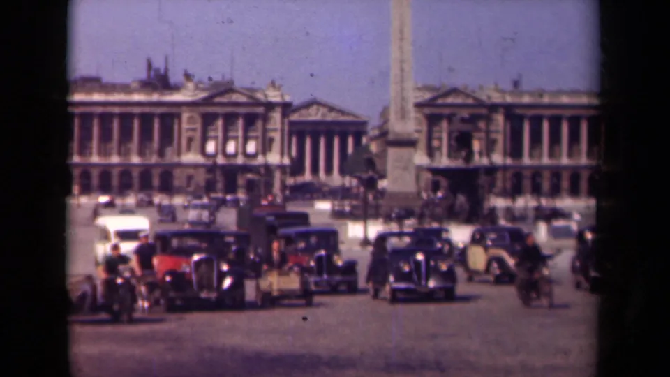 1939 Paris street scene with several dark automobiles, a cyclist, pedestrians, an obelisk, and large neoclassical facades in soft 8mm color.