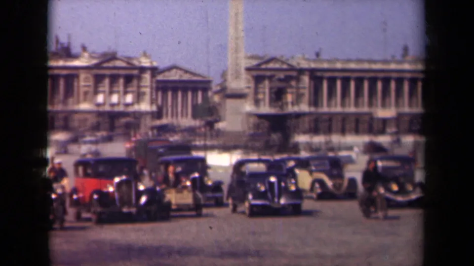 1939 Paris frame showing a red car, dark sedans, cyclists, pedestrians, an obelisk, and a long neoclassical building across the square.