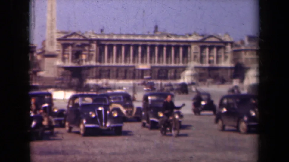 1939 Paris home-movie frame with dark automobiles, riders on bicycles, pedestrians, the obelisk base, and a large neoclassical building in the background.