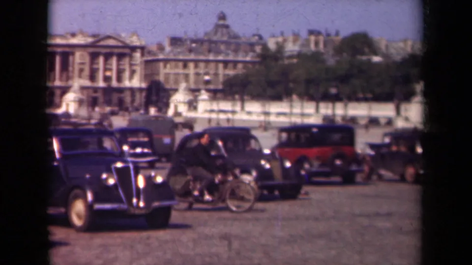1939 Paris street scene with a rider moving through vintage car traffic before formal architecture and trees.