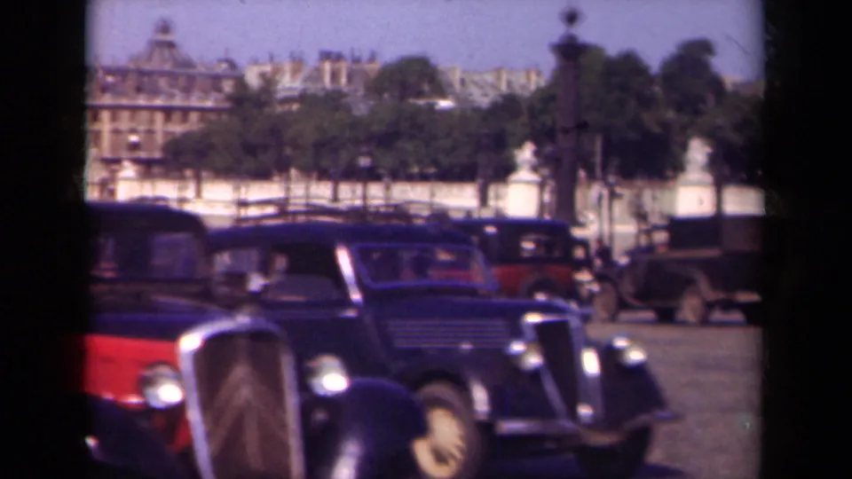 1939 Paris frame with close passing automobiles, a red vehicle edge, distant trees, statues, and formal buildings around the square.