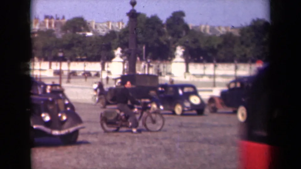 1939 Paris home-movie frame showing a rider, multiple vintage cars, statues, trees, and a tall street feature in a public square.