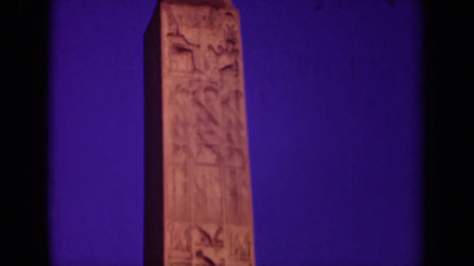 1939 Paris frame showing a carved obelisk section with Egyptian-style figures and vertical marks, rendered in warm stone tones against purple.