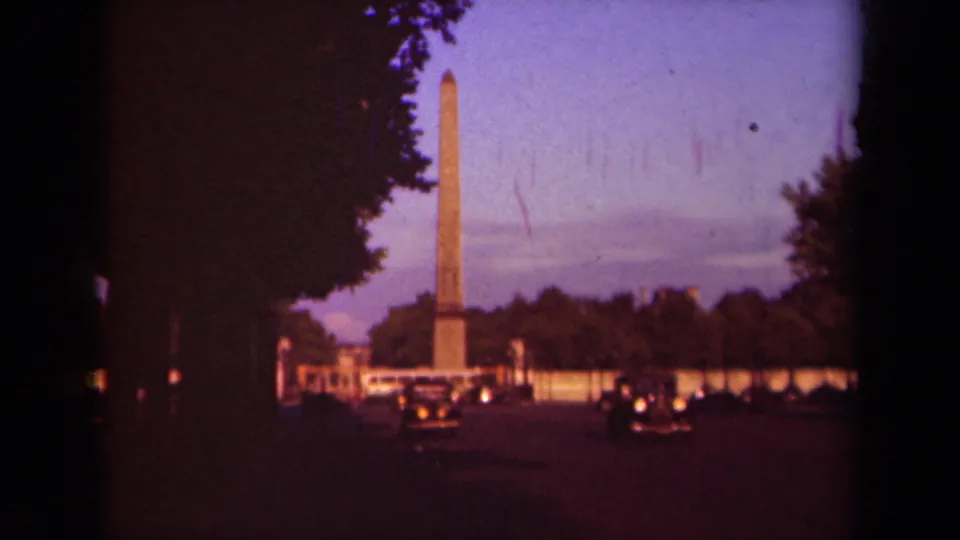Wide Paris home-movie frame with a tall obelisk, shadowed trees, cars on the avenue, and a purple evening sky.