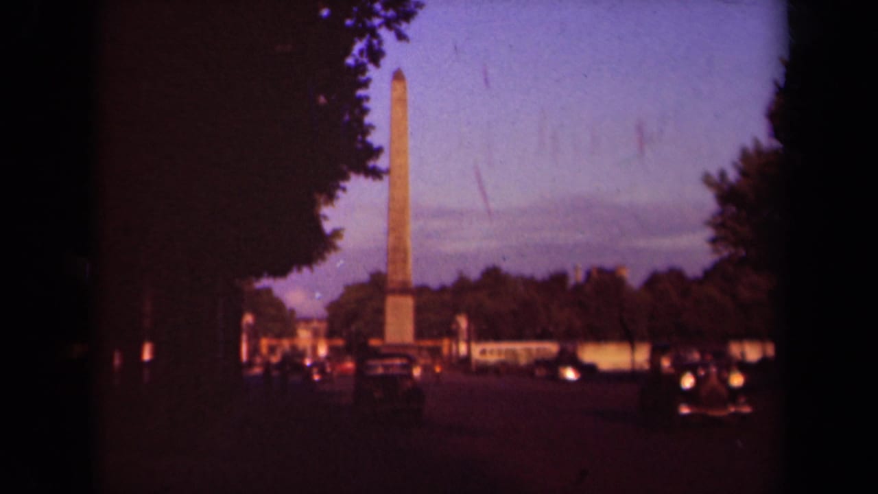 Stone obelisk framed by dark trees and street traffic at dusk.