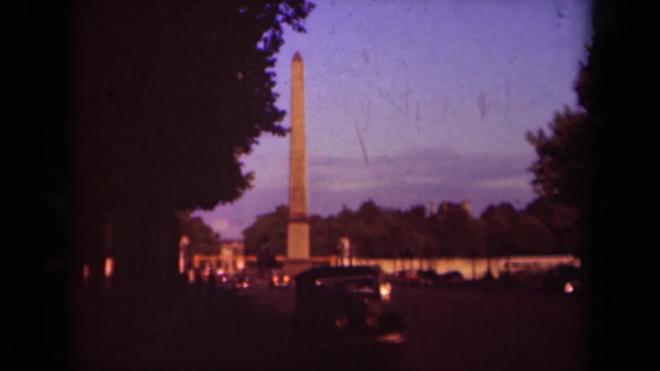 Underexposed 1939 Paris street frame with an obelisk rising behind traffic and trees at dusk.