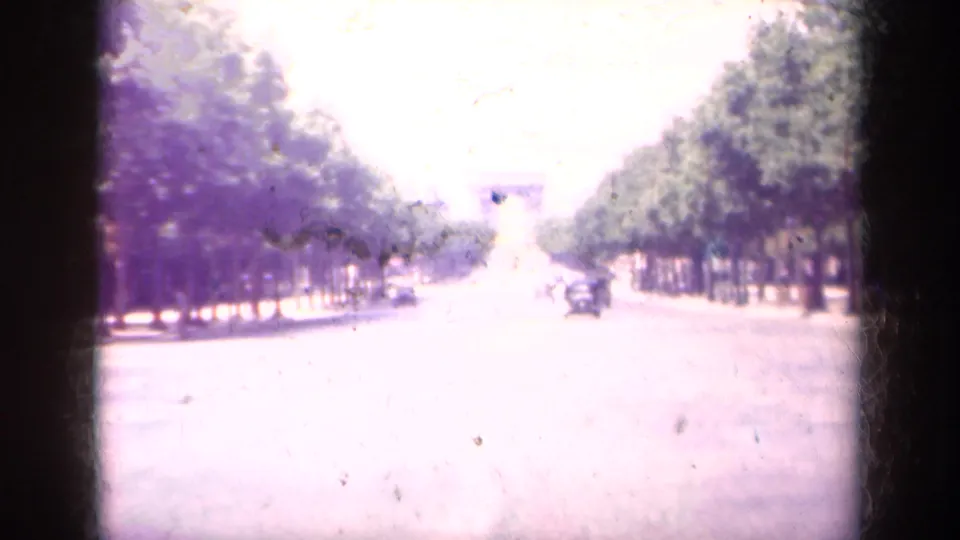 1939 Paris home-movie frame looking down a broad tree-lined boulevard toward a distant triumphal arch, with cars on the roadway.