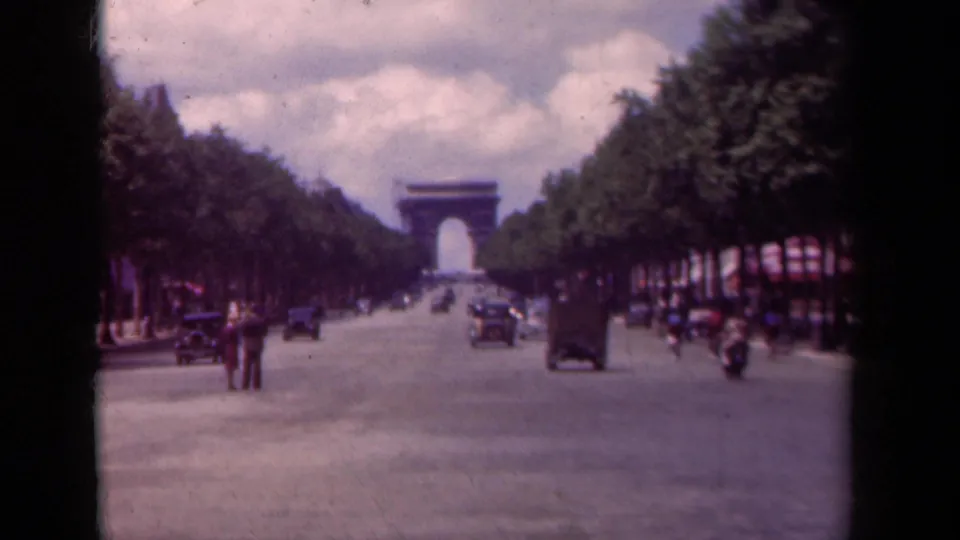 1939 Paris street view toward the Arc de Triomphe, with pedestrians crossing or standing near period automobiles on the Champs-Elysees.