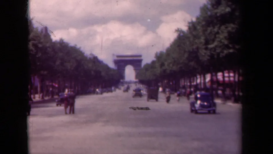 Paris boulevard scene looking toward the Arc de Triomphe, with small groups of pedestrians and period vehicles in motion.
