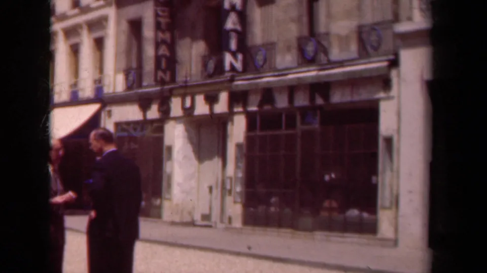 Paris street facade with a theater sign, storefront windows, an awning, and pedestrians partly visible in the foreground.