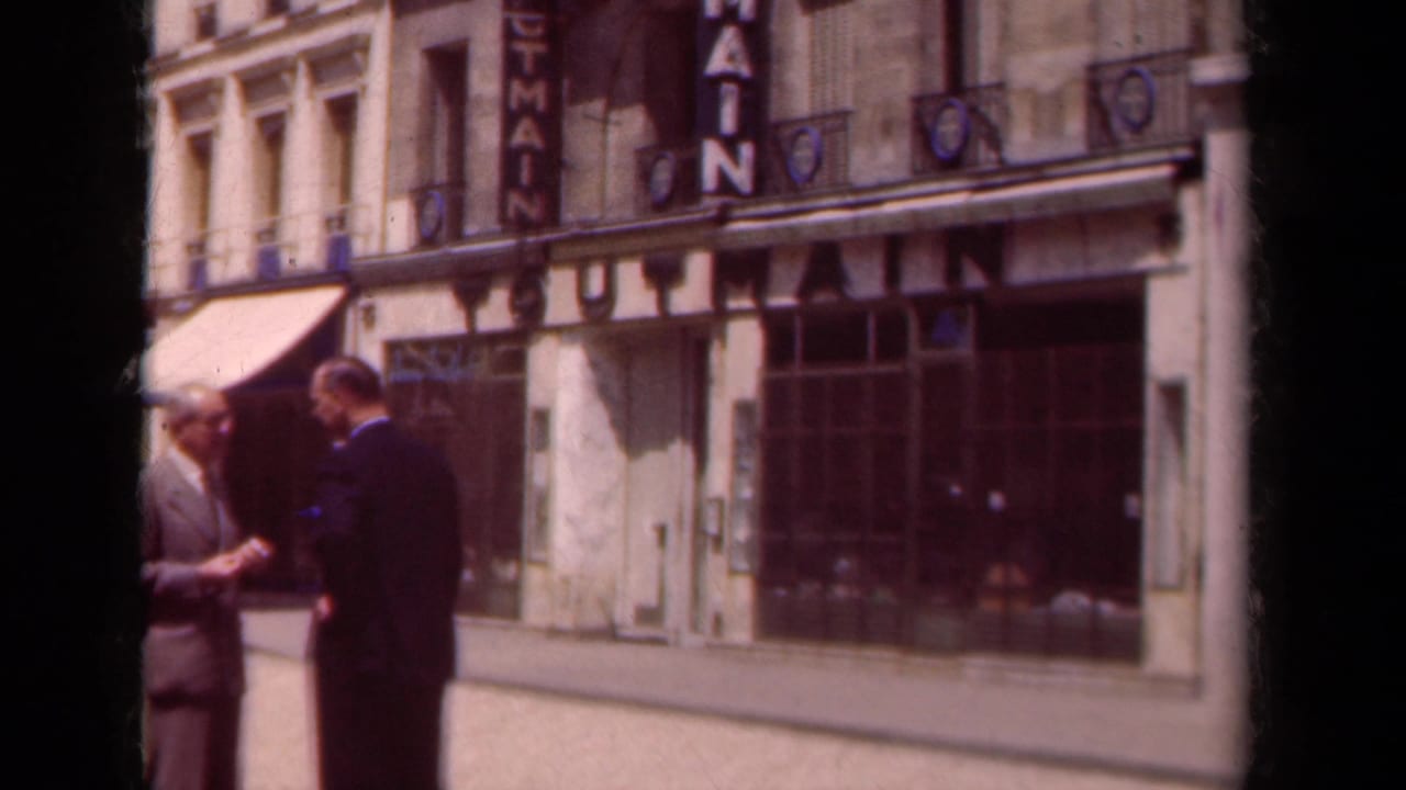 Two suited men stand on a Paris sidewalk in front of awnings and a vertical MAIN sign.