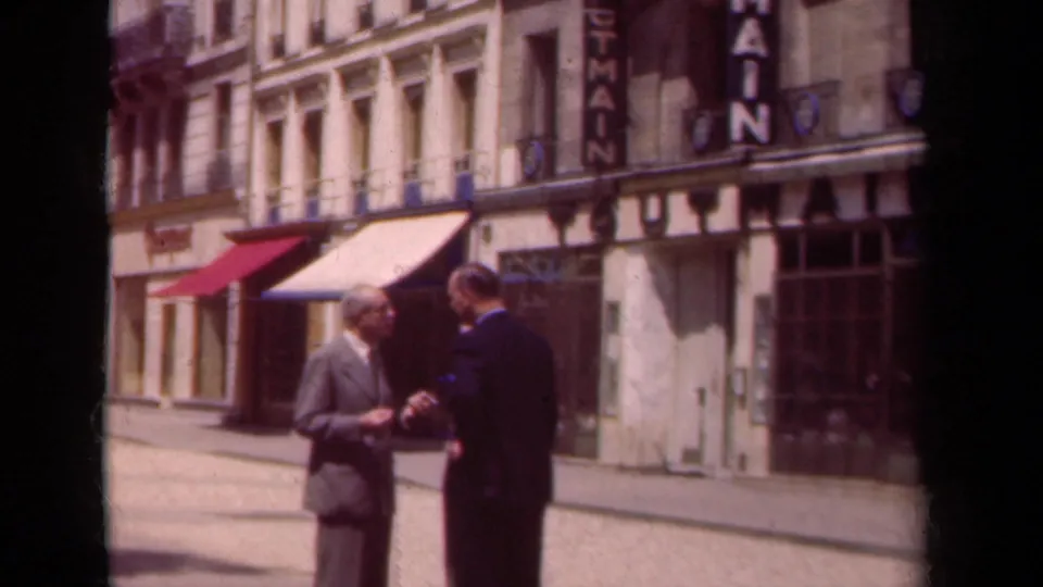Two suited men continue their sidewalk conversation on a 1939 Paris street lined with shopfronts, awnings, and upper-story windows.