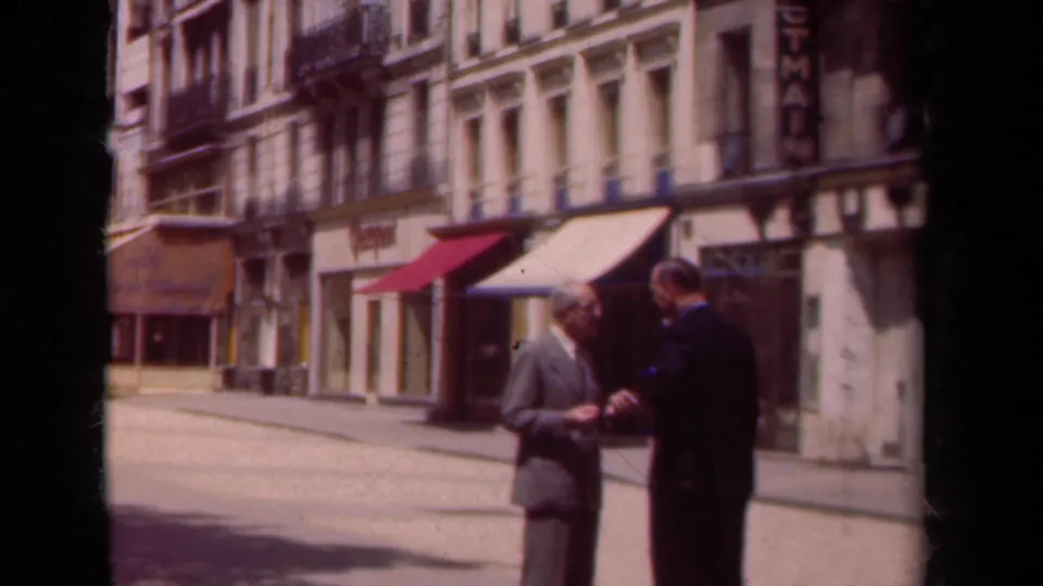 On a Paris sidewalk in 1939, two suited men stand before a row of storefronts with colored awnings and masonry facades.