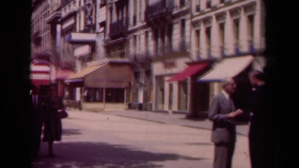 A pedestrian woman passes through the 1939 Paris street view as two suited men converse beside awnings and shopfronts.