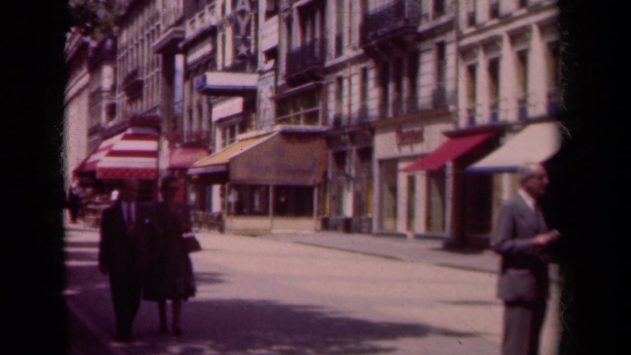 A couple walks along a Paris street lined with storefronts and striped awnings.